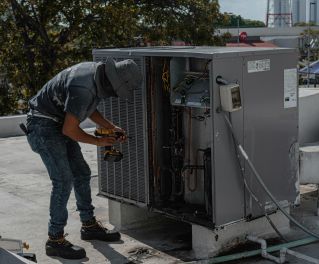 A worker in a bucket hat repairs an outdoor air conditioning unit on a rooftop.